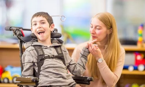 Un enfant polyhandicapé sourit dans une salle de classe près d’une femme adulte.
