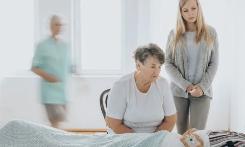 Deux femmes au chevet d&rsquo;un homme allong&eacute; dans son lit d&rsquo;h&ocirc;pital.