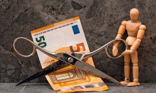 Une figurine (homme) en bois coupe un billet de 50 euros avec d’énormes ciseaux.