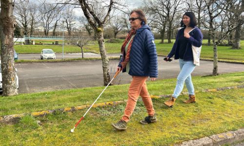 Une femme teste la piste d’essai et marche, avec sa canne blanche, dans l’herbe.