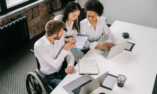 Trois coll&egrave;gues, dont un en fauteuil roulant, discutent dans un bureau.