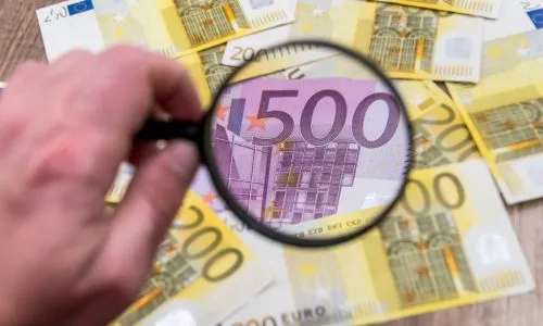 La main d’un homme tenant une loupe au-dessus de billets de 200 et 500 euros.
