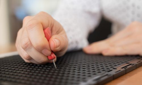 Gros plan sur les mains d’une personne écrivant en braille avec un poinçon.