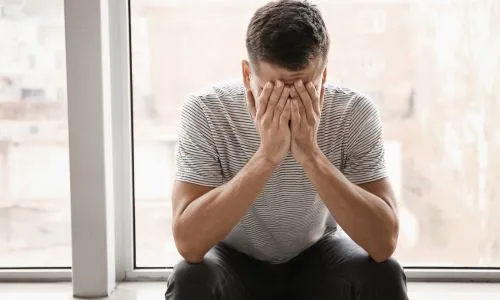 Un homme tient sa tête entre ses mains, près d’une fenêtre.