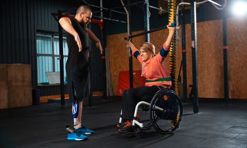 Femme en fauteuil roulant soulève sa barre de musculation face à un homme amputé
