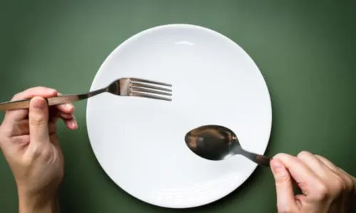 Des mains tienent une fourchette et une cuillère au-dessus d’une assiette vide.