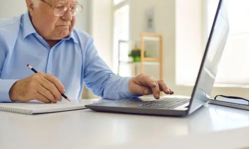 Un senior pianote sur son ordi et prend des notes sur un cahier.