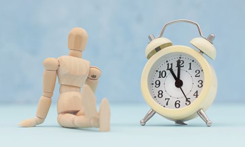 Un petit bonhomme en bois attend les jambes croisées près d’une horloge.