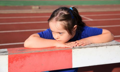 Jeune fille avec trisomie, le regard vide, pose ses bras sur un saut d’obstacle.