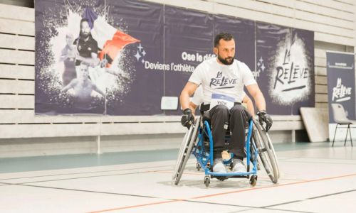 Un homme dans un fauteuil roulant adapté au sport devant l’affiche de La Relève.
