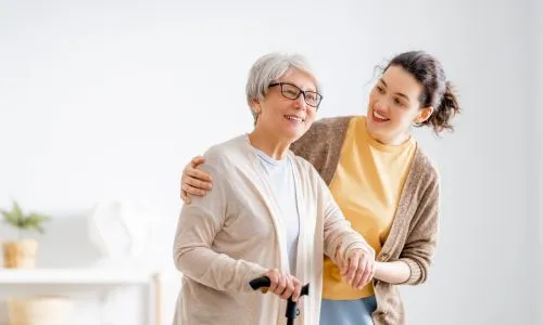 Une jeune femme soutient une femme avec une canne, souriante.