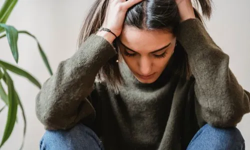 Une jeune femme se tient la tête, assise contre le mur, le regard vers le bas.