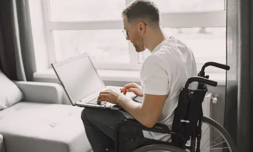 Un homme en fauteuil roulant sur son ordinateur face à la fenêtre.