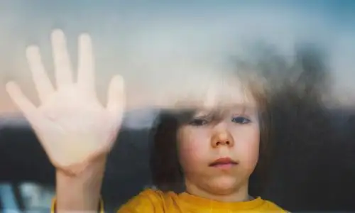 Un enfant triste regarde par la fen&ecirc;tre, sa main appuy&eacute;e contre la vitre.