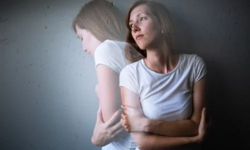 Une femme, l’air triste, les bras fermés, adossée contre un mur grisâtre.