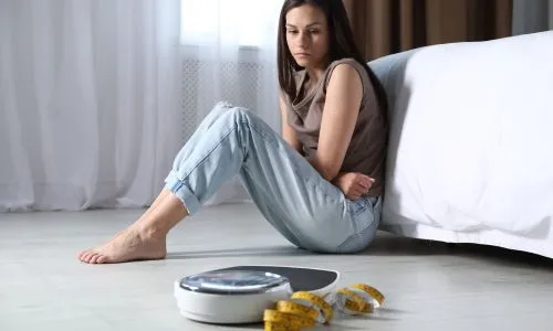 Une jeune femme assise au pied du lit regarde une balance à côté d’un mètre.